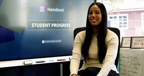 A smiling woman sits in front of a SMART Board displaying 'Handout: Student Progress' on the screen.