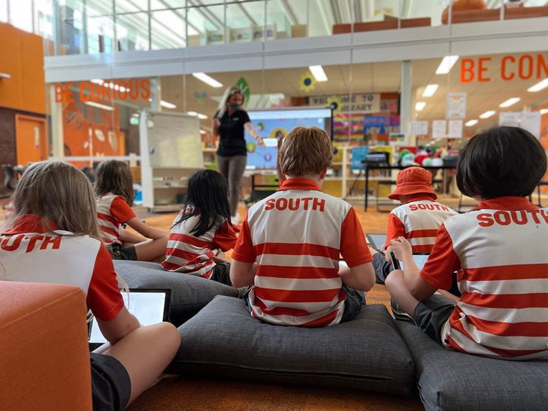 Students in red and white 'South' uniforms sit on cushions in a library while a teacher conducts a lesson on a SMART Board.
