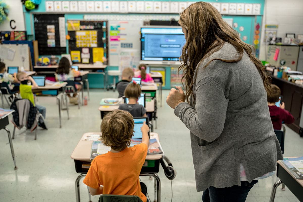 Teacher assisting students in a classroom setting, with children using tablets and a SMART Board in the background.