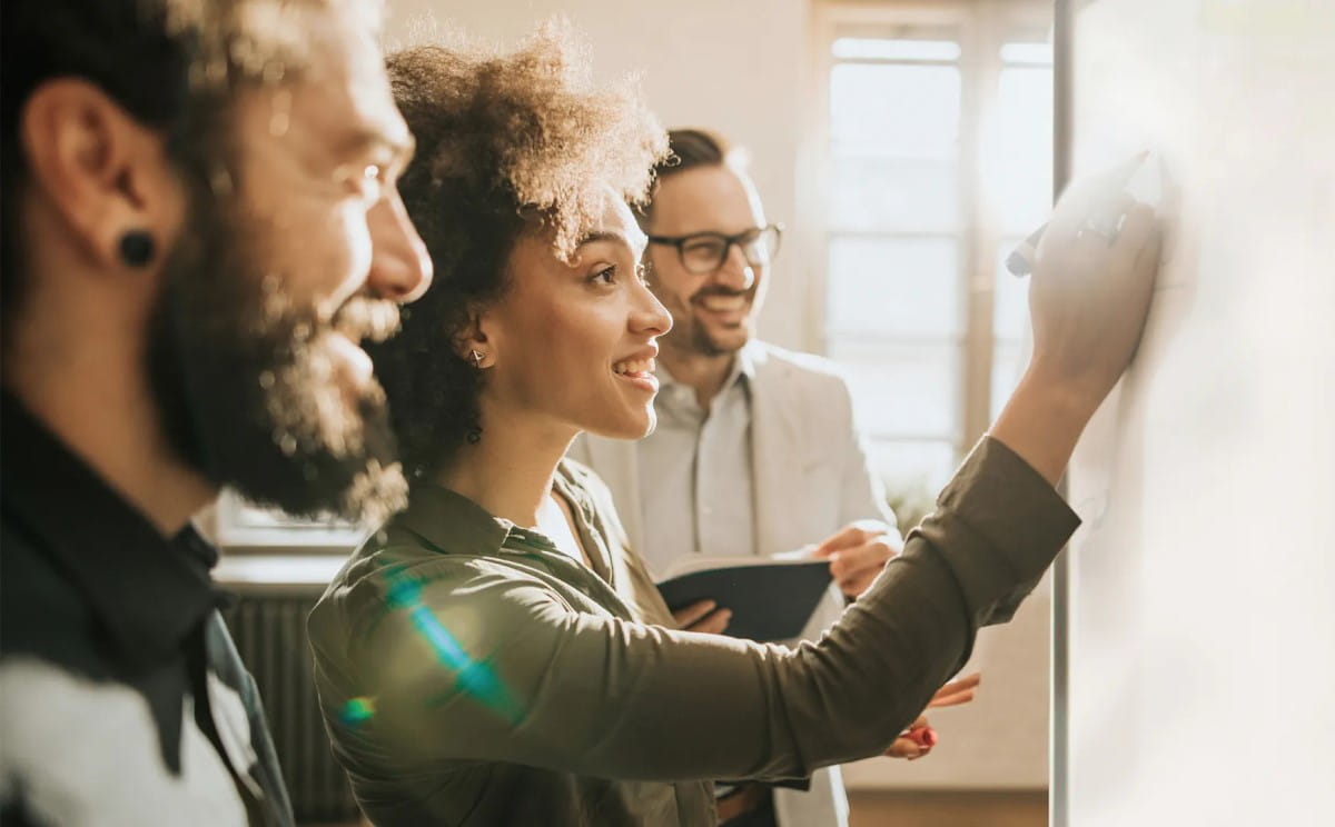 A diverse team engaging in a collaborative session, with a smiling woman writing on an interactive display as her colleagues, two men with one in the background holding a notebook, watch and discuss the content enthusiastically.