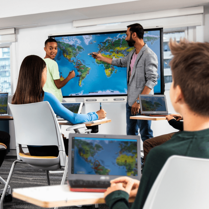 Students in a classroom look on as a teacher explains a topic on a SMART board with a world map.